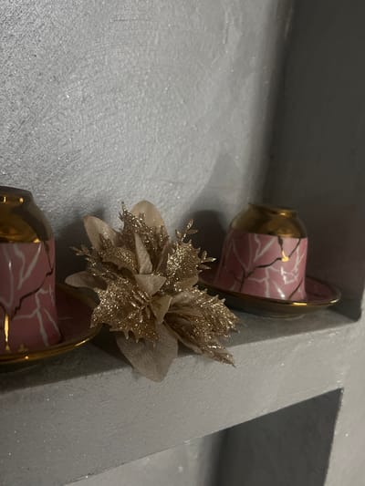 The scene is an interior display within a structure in Vaglen, Bulgaria. A light-colored, textured shelf, exhibiting glitter flecks, serves as the base for three decorative items. On the left side of the shelf, an inverted, small porcelain cup is positioned. This cup is pink with an irregular white and gold crackle-like pattern and a gold rim, resting on a matching pink saucer with a gold rim. A second, identical inverted cup and saucer set is located on the right side of the shelf. Centrally placed between the two cup sets is a single, artificial gold-colored flower, featuring layered fabric petals and spiky leaf-like elements, all heavily coated in glitter. The wall directly behind the shelf is uniformly light-colored and exhibits a textured finish, consistent with the shelf surface. No individuals are present. All objects are static and arranged for display purposes.