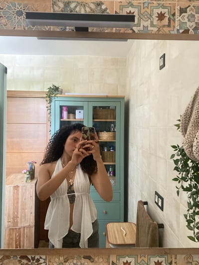This media file is a self-portrait taken in a bathroom setting. The main subject is a young woman with curly dark hair, wearing a white ruffled top and dark pants. She is holding a smartphone in her right hand, capturing her reflection in a large mirror. Her left arm is also visible, with a cross necklace around her neck and a ring on her finger. Her expression is neutral as she takes the photo.

The background of the bathroom is reflected in the mirror. To the left, there is a wall with decorative tiles in geometric patterns of beige, brown, blue, and green. A section of a black object is visible, possibly a piece of furniture or a light fixture. To the right of the subject's reflection, a pale green cabinet with wooden drawers is visible, filled with various items like baskets and bottles. Above the cabinet, a knitted item and green leaves from a plant are reflected, suggesting a decorative element in the room. The ceiling is tiled with square, light-colored tiles. A modern, square light fixture is mounted on the ceiling.

The lighting suggests it is daytime, likely indoors. The overall mood appears casual and personal, characteristic of a private space. There is no visible text in the image. The location cue provided is Zeneta, Spain, which implies the setting is a bathroom in that region.