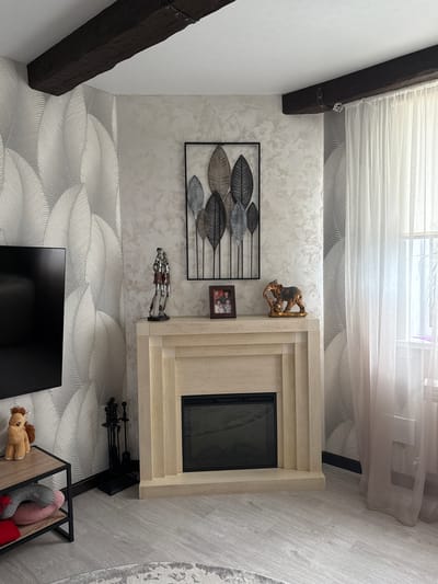 This media file captures an interior scene, likely a living room, with a modern and cozy aesthetic. The room features light-colored walls, some adorned with a subtle textured or patterned wallpaper, and a light wood-look flooring. An exposed dark wooden beam runs across the ceiling and down a wall, adding a rustic touch.
A prominent feature is a light-colored, possibly wooden, fireplace mantel with a dark, modern insert. To the left of the fireplace, on the textured wall, hangs a large black metal wall art piece depicting stylized leaves in varying shades of grey and silver. Below this, a small framed photograph is visible. Above the fireplace mantel, a golden decorative figurine, possibly depicting elephants or monkeys, is perched. Another decorative piece featuring two silver knight-like figures stands on the mantel to the left of the fireplace opening.
Below the fireplace, black metal fireplace tools are neatly stored. To the left, a television screen is visible, angled towards the viewer. A low storage unit with a wooden base and black metal frame is positioned to the right of the TV, holding several plush toys including a brown horse, a striped animal, and a red heart-shaped item.
Natural light filters into the room through a window covered by sheer white curtains, suggesting it is daytime. The flooring to the right of the fireplace is a light wood-look material, and a patterned area rug is partially visible on the far right. The wallpaper has a distinct wavy or leaf-like pattern, creating a sense of movement.
The scene is static, with no activity taking place, and no discernible text is visible on any objects. The setting is an interior space in Krapivka, Russia.