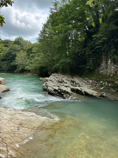 This image depicts a scenic view of a river flowing through a lush, green environment. The river is a vibrant turquoise color, with white rapids forming in the foreground, indicating a swift current. Large, light-colored rocks are scattered along the riverbanks and in the water, some covered with moss. Dense, green foliage and trees line the river, creating a natural, almost wild atmosphere. The sky above is overcast with dramatic clouds.
To the left of the frame, on a rocky outcrop, a person is sitting with their legs extended towards the water, partially visible. Their presence suggests a moment of relaxation or contemplation amidst the natural beauty.
The overall impression is one of tranquility and untouched nature. This scene likely captures a section of the river in the mountainous region surrounding Lebache, Georgia, a location known for its natural beauty and pristine landscapes. The water's clarity and the surrounding vegetation point to a healthy ecosystem.