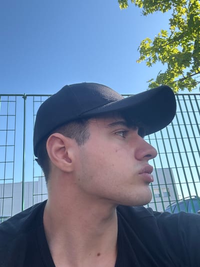 This image, captured in Ancona, Italy, presents a close-up, low-angle perspective of a young adult male looking upwards. The subject is positioned in profile, facing towards the upper left of the frame, with his head and part of his torso visible. He wears a plain black t-shirt and a black baseball cap, worn backwards with the brim pointing upwards. His gaze is directed towards the sky, and his lips are slightly parted.

The scene is set outdoors under a clear, bright blue sky, indicating sunny daytime weather. In the upper left portion of the background, lush green leaves and branches of a tree are visible against the sky, suggesting nearby foliage. A distinctive green wire fence or grid structure extends horizontally across the middle and right side of the background. Behind this fence, on the right, a modern building with light-colored walls and discernible windows can be seen.

The subject's expression appears neutral, perhaps contemplative or relaxed, as he gazes upwards. His skin tone is light, and he has short, dark hair visible around his ear and temple, along with some visible stubble on his jawline and chin. There is no discernible text within the image. The overall composition conveys a tranquil moment spent outdoors, possibly observing the surroundings or enjoying the pleasant weather.