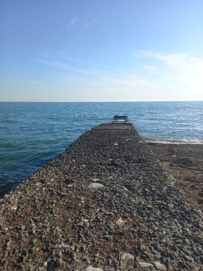 The image depicts a concrete pier jutting out into a calm, blue sea under a bright, partly cloudy sky. The pier is constructed from a rough aggregate of grey and brown stones embedded in concrete. It appears to be a man-made structure, possibly a breakwater or part of a small harbor.
At the end of the pier, a weathered, dark metal structure, possibly a seating area or a small shelter, is visible, partially submerged in the water. The water is a deep blue, with gentle ripples on its surface. The shoreline, to the right of the pier, is also made of pebbles and rocks.
The sky is a gradient of light blue, with wispy white clouds scattered across it. The sun is bright, casting shadows on the pier and illuminating the water. The overall impression is one of a serene, seaside setting on a clear day. The time of day appears to be late morning or early afternoon, given the quality of the light. The location context provided is Gwandra, Abkhazia, suggesting a coastal region. There is no visible text in the image.