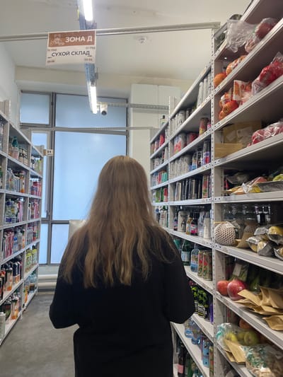 The image depicts the interior of a warehouse or storage facility, possibly a retail stockroom. A woman with blonde hair, seen from the back, is walking down an aisle. She is wearing a black top. The aisle is lined with tall metal shelves filled with various products. On the left side, shelves are stocked with bottles of drinks, juices, and other bottled goods. On the right side, the shelves contain a mix of packaged food items, including fruits like pomegranates and apples, as well as bagged produce and boxed goods. Overhead, there is a sign that reads "ЗОНА Д" (ZONE D) and "СУХОЙ СКЛАД" (DRY WAREHOUSE) with directional arrows indicating rows. The lighting is provided by fluorescent ceiling lights. The overall environment suggests an organized storage area. The city of Imeni Morozova is mentioned, which is a town in the Leningrad Oblast, Russia, known for its industrial and agricultural enterprises, making a warehouse of this nature plausible in that location.