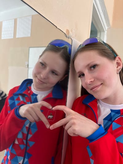 A young woman is pictured resting her head on a reflective surface, seemingly a mirror, which creates a reflection of her face and part of her upper body. She is wearing a red and blue patterned jacket over a white shirt. She also has sunglasses pushed back on her forehead. Her hand is visible, forming a small rectangular shape, revealing an emblem on the jacket that depicts a white bear with a shield. This emblem suggests a connection to the city of Orel, Russia, as it is a part of the city's coat of arms. The background of the reflection shows an interior, possibly a room with window frames.