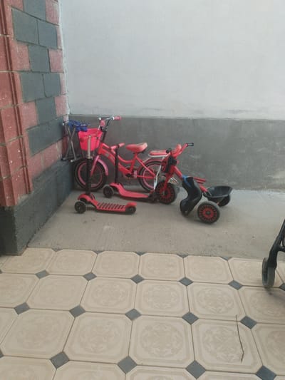 Three children's toys are arranged in a corner next to a brick wall. From left to right, the toys are a red scooter, a pink bicycle with a basket, and a red tricycle with a black seat. The scooter is a two-wheeled model with a pink deck and black handlebars. The bicycle is a bright pink with black handlebars, a pink basket, and training wheels. The tricycle is also pink and red, with a black seat and a black basket. The toys are on a concrete surface with decorative tiles in the foreground. The scene appears to be outdoors, possibly in a yard or patio area. There is no visible text in the image. The lighting suggests it is daytime.
