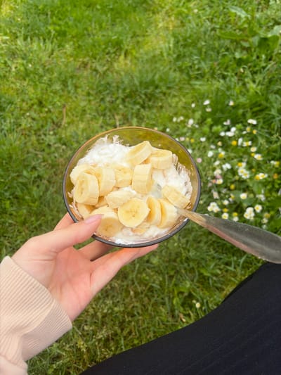 A high-angle shot depicts a hand holding a glass bowl filled with what appears to be cottage cheese and sliced bananas. A spoon is partially inserted into the mixture. The backdrop consists of a green lawn with scattered small white flowers. The lower portion of a person's leg clad in black fabric is visible on the upper right of the frame. The setting appears to be outdoors. There is no information to suggest that the photo was taken in the city of Brusen, Bulgaria.
