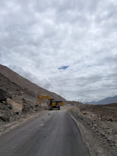 The image depicts a road construction site in Panamik, India. A yellow excavator is positioned on the right side of the road, with its arm extended. The road itself appears to be paved but has some potholes. To the left of the road, a steep, rocky mountainside rises, showing signs of excavation and debris. The landscape is barren and mountainous, with sparse vegetation. The sky is overcast with heavy clouds, suggesting a cloudy or potentially rainy day. In the background, more mountains are visible in the distance, shrouded in haze. There are a few utility poles along the side of the road. A small blue patch of sky is visible amidst the clouds. The scene conveys a sense of ongoing development or repair work in a rugged, remote environment.