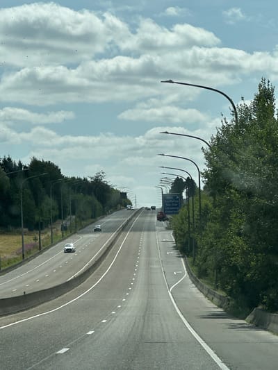 The image shows a highway with multiple lanes, curving into the distance. The road is lined with trees on both sides, and tall lampposts with lights are positioned along the highway. In the sky, there are white, fluffy clouds against a blue backdrop. Several cars are visible on the road, traveling in the same direction. On the right side of the highway, a blue road sign is present, indicating directions to various locations including "Houffalize", "Ste-Ode", "Libramont", and "Amberloup". This sign suggests the location is near Amberloup, Belgium. The environment appears to be a rural or semi-rural area, with dense foliage and grass fields visible beyond the trees on the left. The weather seems to be fair, with visible sunlight casting shadows.