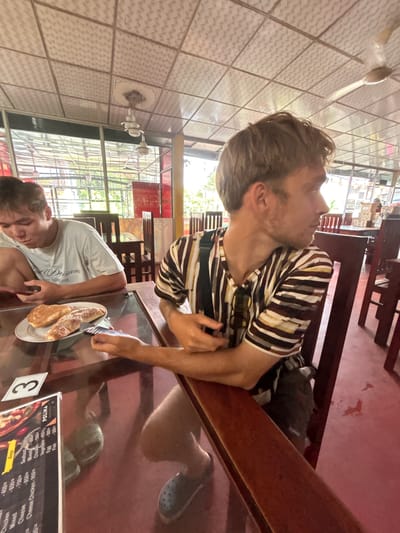 Two young men are seated at a table in an open-air restaurant with a red floor. The man on the left, with dark hair, is wearing a white t-shirt with an inscription and is looking down at a plate of bread rolls. He is holding a fork. The man on the right, with blond hair, is wearing a striped t-shirt and is looking away from the camera, over his right shoulder. He is resting his arm on the table. A menu with food items and prices is visible on the table. The ceiling has a patterned tile design and ceiling fans. The restaurant appears to be located in Walasmulla, Sri Lanka.