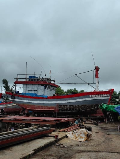 The image displays an outdoor boat repair yard, consistent with a location in Thuận An Ward, Vietnam, under a heavily overcast, grey sky. A large wooden fishing vessel is the central subject, elevated on wooden blocks, indicating it is out of the water for maintenance or construction. The boat's hull features horizontal bands of red (upper), grey (middle), and a darker red/primer (lower). The superstructure is light grey with red accents on the roof edges. The registration number "TTH-92566-TS" is clearly visible in white text on the stern.

The vessel is equipped with multiple masts and poles; a long, slender pole extends forward from the superstructure, likely a fishing boom. An orange flag is visible on a forward mast, and a tattered red fabric or flag is draped from a vertical pole at the stern. Blue tarpaulins cover sections of the deck or cabin roof. A black, circular fender is attached to the port bow.

To the left of the main vessel, parts of additional wooden boats, painted similarly in red and grey, are visible, also dry-docked. The ground in the foreground and midground is an uneven surface composed of concrete and dirt, appearing damp or muddy in areas. Scattered across this ground are various industrial materials and debris: stacks of reddish-brown metal sheets or panels resting on wooden beams, bundles of metal pipes, discarded wooden planks, and a crumpled white and yellow tarp. A red and a white paint can are visible near the stacked materials. In the mid-right, a green tarp covers an A-frame support structure. Green tree foliage is visible in the background, behind the dry-docked vessels. No individuals are present in the scene.