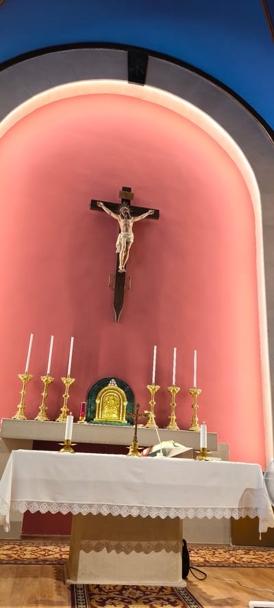 This image depicts an altar in what appears to be a church or chapel. At the center, mounted on a pink wall, is a wooden crucifix with a figure of Jesus. Above the crucifix, a dark emblem is visible. The altar itself is covered with a white cloth, and several golden candlesticks with white candles are arranged on it. To the left, there are seven candlesticks on the altar shelf, with the far left one unlit, and the others appearing to be lit or recently extinguished. A single white candle is lit in a candlestick on the altar cloth in the foreground. Behind the altar, in the center, is a small, ornate golden shrine with a green marble surround. To the right of this shrine, there are three more unlit golden candlesticks. A small brass crucifix stands on the altar cloth to the right of the golden shrine. The floor is wooden, covered in part by a patterned rug. In the upper portion of the image, the ceiling is visible, with sections of blue and what appears to be a curved archway with subtle lighting. The overall scene is static and devotional. No individuals are present in the image. The location of Corró D'avall, Spain, is not visually indicated within the image itself.