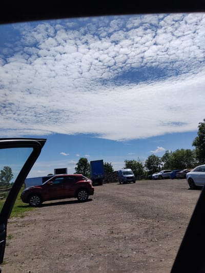 The image, apparently taken from inside a vehicle with the driver's side door open, shows a partially visible parking area on a sunny day. The location is deduced to be in the city of Stafford, United Kingdom. To the right, a gravel-covered lot is observed, featuring several parked vehicles including a red hatchback, a light blue van, and other cars in the distance. A dark shadow partially obscures the right side of the parking area. A few trees dot the edge of the parking lot. To the left, the sky is a mix of blue and white, filled with puffy, cloud formations. The open driver's side door provides a frame for the scene. The interior of the vehicle from which the photo was taken is mostly black. No people are discernible in the image.