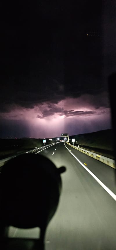 The image captures a nighttime scene on a highway in Malpartida de Plasencia, Spain, during a lightning storm. The highway stretches into the distance, illuminated by streetlights and the white lines marking the lanes. Guardrails line the sides of the road, reflecting the light. In the foreground, the dark silhouette of a vehicle's interior, possibly a dashboard or windshield, is visible, obscuring a portion of the view. The sky is filled with dark, heavy storm clouds, punctuated by bright flashes of lightning that cast a purplish glow on the clouds and the landscape. Distant lights, possibly from other vehicles or settlements, are visible on the horizon. The overall impression is one of dramatic weather occurring while traveling on an open road.