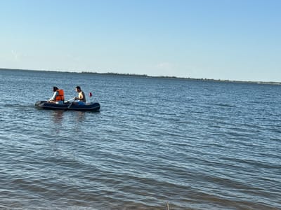 Two individuals are visible in a dark inflatable boat on a calm body of water under a clear, bright blue sky. The person in the front of the boat is wearing an orange life vest and facing away from the camera, while the person in the back is bare-chested, facing forward, and holding an oar as if paddling. A small red buoy or marker is floating in the water behind the boat.
The scene depicts a recreational activity, likely boating or rowing on a lake or large river. The water exhibits gentle ripples, reflecting the clear sky. In the distance, a low, tree-lined shoreline stretches across the horizon, suggesting a natural landscape. The overall impression is one of a peaceful, sunny day spent outdoors. The location context indicates this is in Zerendi, Kazakhstan, known for its lakes. There is no visible text in the image.