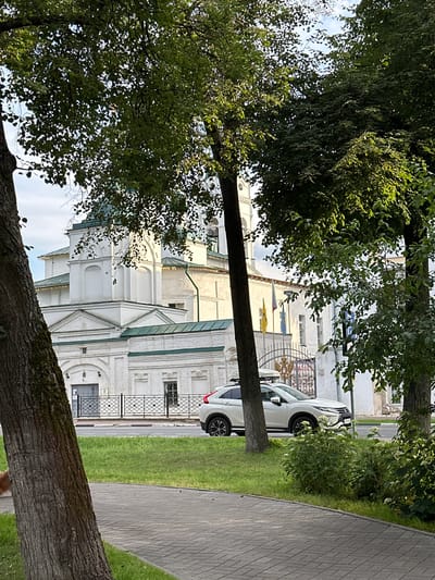 A white SUV with a roof rack is parked on the side of a road next to a paved walkway and a grassy area. In the background, a white building with green trim and a green dome is visible. Several trees with green foliage are in the foreground, obscuring parts of the building. The sky is partly cloudy. The location is Putyatino (Semivragovskaya S/A), Russia.