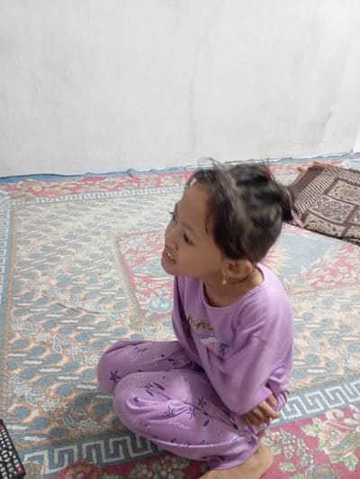 An individual, identified as a young female of apparent Southeast Asian ethnicity, is depicted seated on a patterned rug within an interior space in Bulukandang, Indonesia. The individual has dark hair tied back and is wearing a long-sleeved light purple shirt and matching long pants adorned with white star patterns. A small golden hoop earring is visible on her right ear. Her head is turned slightly to the left, and her gaze is directed upwards and to the left of the frame; her mouth is slightly open. She is barefoot, and her hands are positioned in her lap. The rug beneath her features complex floral and geometric patterns in red, blue, and cream tones. The background consists of a plain white wall exhibiting minor discoloration. A dark brown cushion with a geometric pattern is visible on the rug behind the individual's right side, and a black object resembling a television remote control is partially visible in the lower-left foreground.