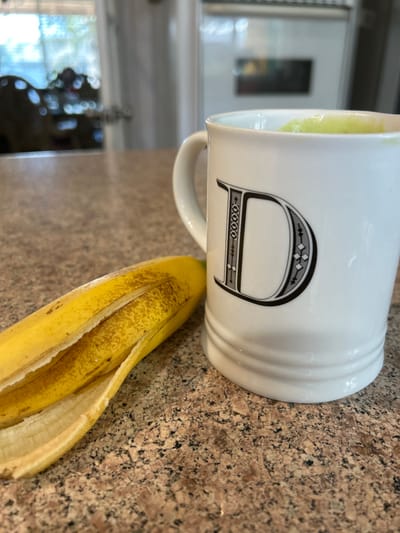A partially peeled, ripe yellow banana, showing brown speckles on its skin and exposed fruit, lies horizontally on a speckled brown and beige granite countertop. Adjacent to the banana is a white ceramic mug featuring a black ornate capital letter 'D' on its side and horizontal ridging near its base. The mug contains a light green liquid, consistent with a smoothie. The background is blurred, indicating an indoor setting, likely a kitchen, with a window displaying indistinct outdoor foliage on the left and a large white appliance on the right. No individuals are visible. This scene is situated in Thousand Oaks, United States.