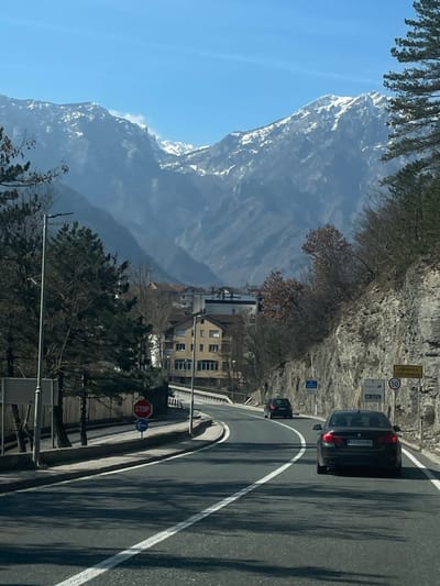 The image depicts a daytime scene in Jablanica, Bosnia and Herzegovina, specifically along a two-lane asphalt road curving slightly to the left. In the foreground, a dark-colored sedan, a BMW with a license plate partially visible as "T09-M-110", travels away from the viewer in the right lane. Further ahead in the left lane, a dark-colored hatchback or small SUV also moves away from the viewer.
To the right of the road, a steep, rocky cliff face with sparse, bare deciduous trees and evergreen shrubs ascends. Road signs are affixed to the cliff face: a yellow rectangular sign reading "Jablanica Јабланица" above a circular 50 km/h speed limit sign, followed by a yellow "PLAC" sign with a right-pointing arrow. Below these are blue rectangular highway signs indicating "E73" and "M17", and another blue sign with an arrow pointing to "Mostar".
To the left of the road, a metal guardrail or fence runs parallel to the asphalt, behind which are several evergreen trees and bare deciduous trees. A street light pole is visible on the left. Further down the road, past the trees, a red octagonal "STOP" sign is posted, accompanied by a blue circular sign featuring a white left-pointing arrow. Behind these signs and trees, multi-story buildings belonging to the town of Jablanica are visible, including a prominent light-colored building with multiple windows and a reddish-brown roof.
In the background, under a clear blue sky, majestic mountains with visible snow patches on their peaks dominate the horizon, creating a mountainous valley setting for the town. The overall lighting suggests bright sunlight, casting shadows on the road and structures.