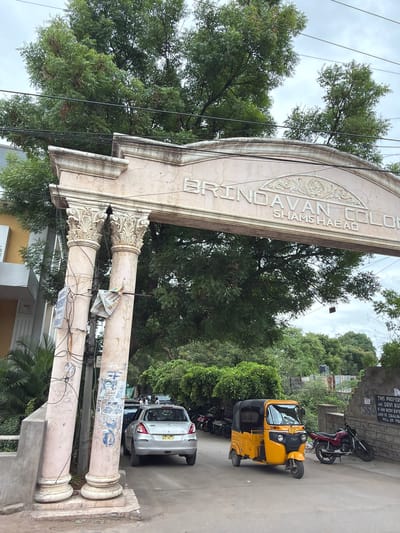 The image captures the entrance archway to "BRINDAVAN COLONY SHAMS HABAD" in Shamshabad, India. The archway, made of stone, is supported by two ornate pillars. Wires and posters are attached to the pillars. Beyond the archway, a silver hatchback car is parked on the left, and a yellow auto-rickshaw is visible in the center. A red motorcycle is parked to the right of the auto-rickshaw, leaning against a stone wall. Several other motorcycles are parked further down the road on the left, behind the car. Lush green trees form a canopy over the scene, with a cloudy sky in the background. A sign is affixed to the stone wall on the right. The overall scene depicts a residential colony entrance with vehicles and foliage typical of an urban Indian setting.