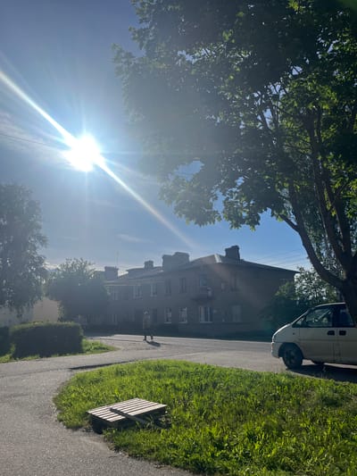 The image depicts an outdoor scene in a residential area, likely in Серебрянский, Russia. A bright sun, positioned low in the sky, casts lens flare across the frame, indicating it's either early morning or late afternoon. The sky is a clear blue, suggesting fair weather.
The foreground features a paved pathway bordered by lush green grass and a wooden bench. To the left, the branches of a large deciduous tree with vibrant green leaves create a canopy, partially obscuring the sky. Beyond the tree, a multi-story brick apartment building is visible. The building has several windows and chimneys, hinting at older architecture.
A white van is parked on the left side of the pathway, close to the apartment building. A solitary figure, appearing to be a person, is walking on the pathway in the middle distance, casting a shadow due to the strong sunlight. The overall atmosphere is serene and peaceful, conveying a sense of a typical day in a neighborhood. The presence of trees, a residential building, and a pathway suggest an urban or suburban setting. No visible text is present in the image.