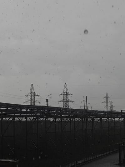 The image shows an industrial landscape under a cloudy, overcast sky, with what appears to be precipitation falling. In the background, several tall electrical pylons are visible, silhouetted against the sky. In the foreground, a dark, multi-tiered industrial structure runs horizontally across the frame, featuring numerous metal supports and horizontal platforms. Barbed wire is visible atop a fence or wall behind the structure. The overall scene suggests an active or formerly active industrial zone, possibly a power substation or manufacturing facility, located in the vicinity of Дүңгілағаш, Kazakhstan. There are no people or animals visible in the image.