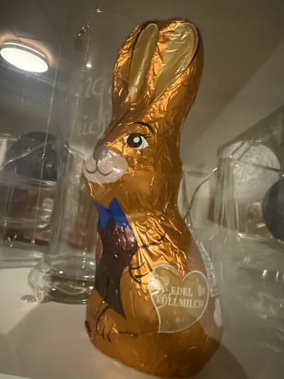 The image shows a close-up, horizontally oriented, eye-level shot of a hollow chocolate Easter bunny wrapped in shimmering gold foil. The bunny is lying on its side, with its head and long ears facing the left of the frame. Its face has a printed expression with large, round eyes and a small, smiling mouth. A blue bow is visible around its neck, and its back is partly obscured by reflective packaging.

On the bunny's body, a heart-shaped label is visible with text printed in a light gold color. The text reads: "EDEL VOLLMILCH".

The bunny appears to be inside a clear plastic container, possibly for display or packaging. The background is softly lit and out of focus, with hints of other objects and reflective surfaces. One such object appears to be a white, woven material. A bright, oval-shaped light source is visible in the lower left quadrant of the image, reflecting off the plastic.

The scene is likely indoors, given the artificial lighting and the presentation of the product. The overall impression is that of a festive confectionery item, commonly associated with Easter celebrations. No specific time of day or weather conditions are discernible. The location context provided is Hiddenhausen, Germany, which suggests the product might be locally sourced or marketed.