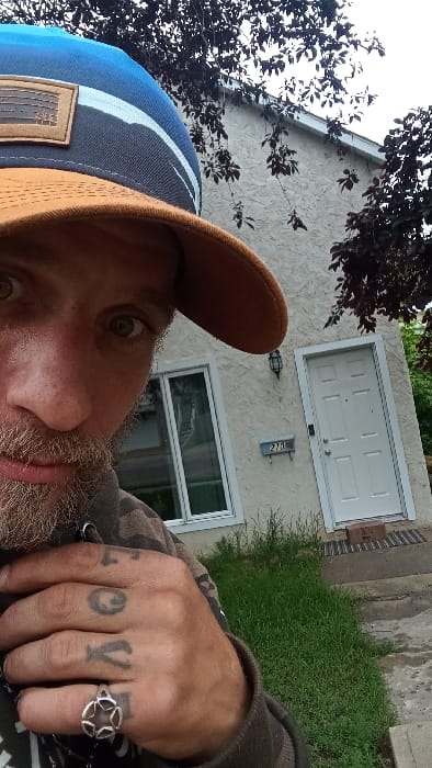 The image displays a male subject in a close-up, upward-angle selfie, positioned outdoors in front of a residential building identified by the municipal address "270" in Medicine Hat, Canada.
The subject's face, visible from the right eye to the nose and mouth, shows light-colored eyes, a light brown beard, and stubble. He is wearing a two-toned baseball cap with a blue crown, a brown brim, and a rectangular leather-like patch on the front. His attire includes a dark, possibly camouflage-patterned long-sleeved garment. The subject's right hand is prominent in the foreground, displaying knuckle tattoos that spell "LOVE" and a silver-colored ring with a five-point star design on the ring finger.
The background consists of a single-story or lower unit of a light beige, stucco-textured building with white trim. A white six-panel entry door is visible on the right, with a dark rectangular doormat and a small brown cardboard box resting on the concrete stoop. To the left of the door, a black, wall-mounted lantern-style light fixture is installed above a light blue rectangular plaque displaying the numerals "270." A large, white-framed window with reflective glass is situated to the left of the entrance. The ground immediately in front of the structure is covered in green grass of varying height, and a concrete pathway is visible in the foreground. Dark-leaved deciduous trees or shrubs partially obscure the building's upper and side portions. The sky above is uniformly overcast, indicating diffused natural light conditions.