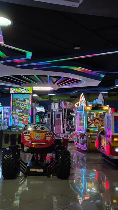 This is an indoor photograph of an arcade in Tlaquepaque, Mexico, likely located within a shopping center or entertainment complex. The scene is dimly lit, with vibrant neon and LED lights illuminating various arcade machines. In the foreground, a red monster truck-style arcade ride designed to resemble a cartoon character with large wheels dominates the lower half of the image. Behind it, several other arcade games are visible, including a racing game with a "Crossy Road" display, a claw machine, and what appears to be a "Walking Dead" themed game. To the right, a yellow and red arcade machine with characters from "Toy Story" is prominent, and further right, another brightly lit ride, possibly a fire truck, is partially visible. The ceiling features a modern design with black panels and colorful, segmented light fixtures that cast geometric reflections on the glossy tiled floor. The overall impression is one of a lively, modern entertainment venue filled with interactive games and stimulating visual elements.