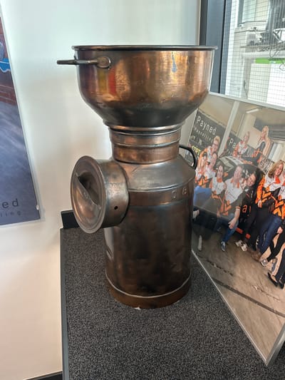 The image shows a large, antique-looking copper container, possibly a milk churn or a similar piece of equipment, positioned indoors on a dark gray carpeted surface. The container is tall and cylindrical, with a wide opening at the top and a handle on the left side. There's a circular opening with a flap on the side of the lower section. Behind the container, to the right, is a large framed photograph or print. This photograph displays a group of people, mostly women, wearing orange and black jackets with "Payned" and "Payroll" branding. They appear to be posing together, some in front of a car. The background of the photograph seems to be an outdoor setting. To the left of the copper container, a portion of a different poster or print is visible, featuring text and a blue/gray background. The environment suggests an indoor commercial or office space, with a light-colored wall behind the container. The overall impression is a display setting, possibly in a company's reception area or exhibition space, showcasing both a historical artifact and promotional material.