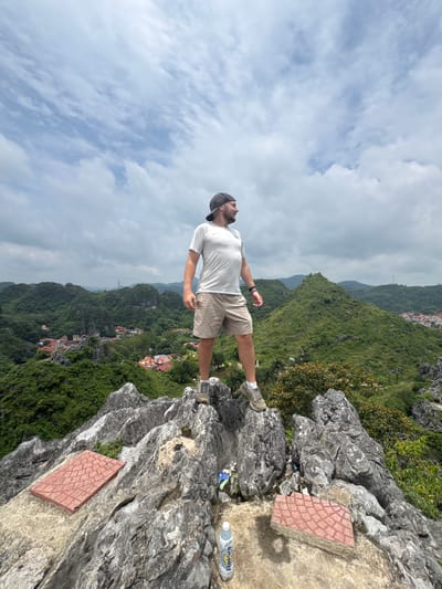 A man is standing on a rocky cliff edge, with his body tilted to the left and his legs spread apart. He is wearing a white t-shirt, khaki shorts, a grey baseball cap, and sneakers. His head is tilted up, looking towards the sky.

The scene is a lush, green mountainous landscape with a village nestled in the valley below. The sky is cloudy, with patches of blue visible. The time of day appears to be daytime, given the natural light. The rocks are grey and uneven, with some vegetation growing between them. On the rocky surface, there are two red tiles with a diamond pattern, and a partially visible bottle of "Revive" water. Some trash is also scattered on the ground near the rocks.

The overall impression is of a person enjoying a scenic view from a precarious vantage point, possibly during a hike or a visit to a viewpoint. The man's posture suggests confidence and a sense of awe.