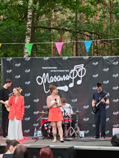 An outdoor live music performance is taking place on a stage in a wooded area, possibly within or near the Ракітніцкі сельскі Савет, Belarus. A black backdrop with a white logo and text "ТВОРЧЕСКОЕ ПРОСТРАНСТВО Маганіф" is behind the performers. The stage is set up on a raised platform, with speakers visible at the front.
Four individuals are actively participating in the performance. On the left, a male is playing a light-colored electric guitar and singing into a microphone. Next to him, a female singer in a bright orange blazer and white dress is holding a microphone and appears to be singing. In the center-rear, another male is seated behind a red drum set. To the right, a male is playing a black electric guitar.
Several other people are visible in the foreground, seated and facing the stage, suggesting an audience. The background consists of tall trees, indicative of a forest or park setting. Colorful triangular flags are strung across the top of the backdrop, adding a festive touch to the event.