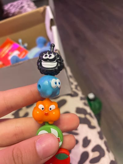 The image shows a close-up of a hand holding a string of small, colorful, toy characters. From top to bottom, the characters are: a black, fuzzy creature with large white eyes and a wide grin; a light blue, round creature with two large blue eyes and a small hole on its side; an orange, puffy creature with large white eyes, an orange nose, and rosy cheeks, which appears to be attached to a green circular object with white markings; and a portion of a red object, possibly another character, at the bottom. The background is out of focus and appears to be a room with a brown wooden floor and a patterned surface, possibly fabric, with a partially visible cardboard box containing some items in the upper left corner. The lighting is artificial and casts shadows. There is no information or visual evidence within the image that can be used to determine the location in Vaganovo Massiv, Russia.