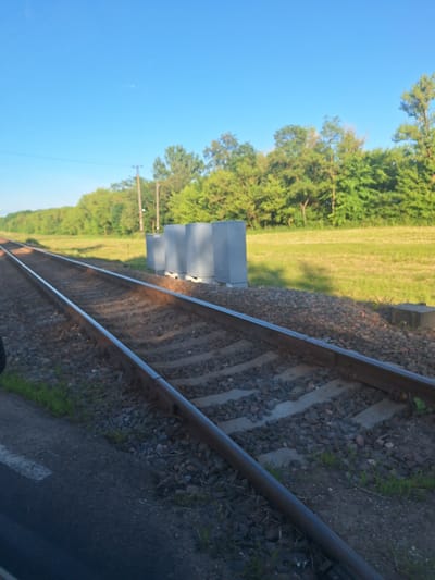 This is a wide-angle, eye-level shot of a railway track running diagonally from the lower left to the upper right of the frame. The sky is clear and blue, with a few faint clouds visible. The railway tracks are laid on a bed of gravel and are bordered by green grass on the right. In the middle ground, there are several tall, rectangular concrete structures lined up next to the tracks. Beyond the tracks and grass, a dense line of trees forms a green backdrop. The scene appears to be in a rural or semi-rural area, possibly near Цімкавіцкі сельскі Савет, Belarus, given the presence of railway infrastructure and natural vegetation. The lighting suggests it is either early morning or late afternoon, with long shadows cast across the gravel. No people or vehicles are visible in the image.