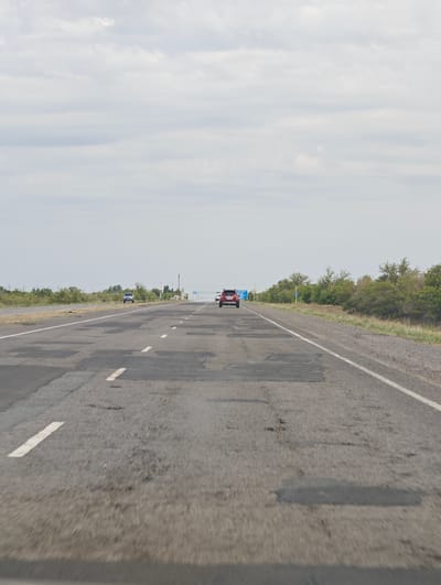 The image depicts a forward-facing view from a vehicle traveling on a highway. The road surface is visibly damaged with numerous patches and potholes, indicating a state of disrepair. White lane markings are present, delineating the driving lanes.
In the distance, several vehicles are visible on the highway. A dark-colored SUV is in the right lane, and further ahead, a red car is in the left lane, appearing to be followed by another vehicle. A lighter colored SUV is in the left lane further down the road.
The setting is a rural or semi-rural landscape with vegetation lining the sides of the highway. Trees and bushes are present on the right, and a less dense line of vegetation is on the left. Utility poles are visible in the distance.
The sky is overcast with a significant amount of clouds, suggesting a cloudy or possibly drizzly day. The lighting is diffused, consistent with an overcast sky, and there are no strong shadows. The time of day is not explicitly indicated but appears to be daytime, likely mid-morning or afternoon.
A blue sign with white text and a speed limit indicator of "60" is visible on the right side of the road. Another sign with blue and white text is further in the distance on the right.
The overall impression is of travel on a highway in Kazakhstan, characterized by a damaged road surface and a cloudy sky. There are no people clearly visible within the frame. The activity is vehicular travel.