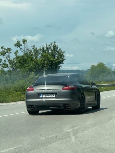 This image captures the rear-left view of a gray Porsche Panamera GTS on the side of a road in Momin Sbor, Bulgaria, under bright daylight. The car is oriented facing away from the viewer, with its prominent taillights, quad exhaust pipes, and black multi-spoke wheels visible. The Bulgarian license plate, "BT 8700 KK", along with the "BG" country code sticker, is clearly affixed to the rear. The model name "PORSCHE" and "Panamera GTS" are embossed on the trunk lid.

The asphalt road, marked by a white solid line, extends from the foreground. Shadows of the car and a metal guardrail on the left indicate a sunny, daytime setting with scattered white clouds in the light blue sky. Beyond the road and guardrail, the scene is dominated by a lush, green landscape, featuring dense trees and bushes that transition into a forested hillside or mountains in the background. The overall setting depicts a scenic, open road environment.