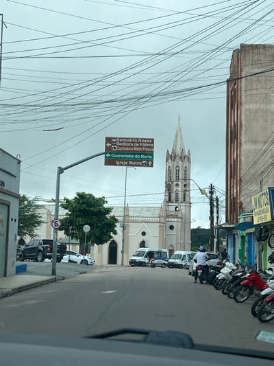 The image depicts a daytime street scene in São Benedito, Brazil, under an overcast sky. A prominent white church, characteristic of neo-Gothic architecture with a tall, multi-tiered spire and arched windows, stands centrally in the frame.
Numerous electrical wires crisscross the upper left portion of the image, extending from a utility pole. On the right, a paved street is lined with parked vehicles. A row of motorcycles is visible on the immediate right, followed by several white vans and other cars further down the street. One person is partially visible near a white van. Commercial buildings with various storefronts line the right side of the street, one showing a partial yellow sign that reads "JM MOTOS."
A brown directional sign on a pole is strategically placed, with white text indicating:
* "Santuário Nossa Senhora de Fátima" (arrow left/straight)
* "Cachoeira Mata Fresca" (arrow left/straight)
* "Guaraciaba do Norte" (arrow right)
* "Igreja Matriz" (arrow right), likely referring to the visible church.
Below this, a red circular sign with a white 'E' crossed out indicates a "No Parking" restriction. A small green tree is situated near the church, and another larger tree is visible further down the street. The scene captures a typical urban setting with a mix of religious architecture, commercial activity, and infrastructure.