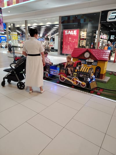 In a shopping mall in Wołomin, Poland, a woman stands near a baby stroller, facing away from the camera. She wears a long, white eyelet dress with a black belt and white sneakers. A young child, possibly a girl, sits in the front car of a miniature train ride. The train, painted black and yellow with red accents and blue lights, is on a track laid out on artificial grass. Behind the train is a small, red-roofed station building with a clock. Another child, a boy wearing a striped shirt and pants, is seated in the train's engine. In the background, storefronts with displays are visible through large glass windows. The floor of the mall is tiled in a light beige color.