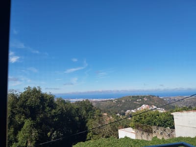 This media file depicts a scenic view from a high vantage point overlooking a coastal landscape in Arucas, Spain. The foreground is dominated by lush green foliage and a partial view of a white building with a decorative concrete wall. The middle ground reveals rolling hills dotted with houses and trees, leading to a distant view of the ocean and a city or town along the coast. The sky is a vibrant blue with a few wispy white clouds.
The overall scene suggests a daytime setting, likely late morning or early afternoon, given the bright sunlight and clear blue sky, indicating good weather. The location cues, such as the architecture of the building and the type of vegetation, are consistent with a Mediterranean or subtropical climate. There are no people or discernible activities taking place in the image; it is a static landscape shot. Notable details include the distant view of what appears to be a port or harbor area with ships, and a hill topped with multiple antenna structures. The image is framed by a dark, vertical element on the left, possibly part of a window frame or doorway. No text is visible in the media file.