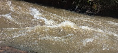 The image displays a turbulent, muddy brown waterway, identified as being within Huruma ward, Kenya. The water is opaque and khaki-colored, characterized by prominent white frothy waves and streaks indicative of a strong current and rapids. Numerous small dark specks are visible throughout the water, consistent with suspended particles or minor debris. The right bank, visible in the upper portion of the frame, is dark-toned and features sparse, dry-looking vegetation, with several pieces of light-colored debris entangled near the waterline. A section of dark reddish-brown earth or rock constitutes the immediate bank in the bottom-left foreground. No individuals or man-made structures are present in the frame.