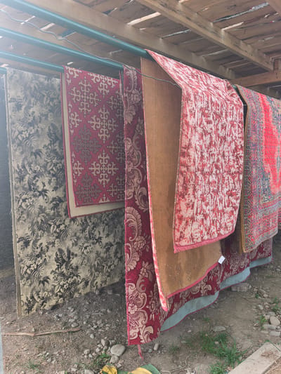The scene depicts multiple rugs hung on a green metal clothesline underneath a wooden structure, possibly a shed or open shelter. The rugs vary in color and pattern, ranging from floral designs in gray and black to geometric shapes in red and beige. The undersides of some rugs are visible, revealing the backing material. The ground is uneven and appears to be a mix of dirt, gravel, and some sparse vegetation. There is no indication of the location being in Semenovka, Kyrgyzstan. No people are visible.