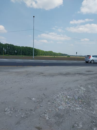 A silver hatchback is parked on the right side of a two-lane asphalt road. The road has white lane markings and a yellow dashed line on the right edge. The foreground consists of a dirt and gravel shoulder with scattered debris. In the background, a dense line of green trees borders a field. A tall, silver utility pole with a small dark box on top stands on the left side of the road, and a traffic sign with black and yellow stripes is visible further down. The sky is blue with scattered white clouds. The overall scene appears to be an outdoor, roadside environment in a rural or suburban area.