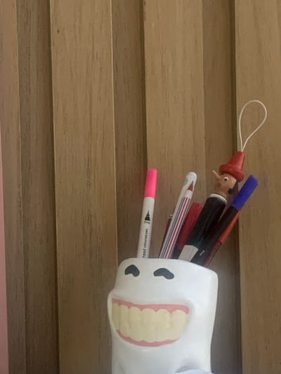 This image depicts a static indoor scene, presumed to be located in Lauro de Freitas, Brazil. The primary subject is a white, ceramic pen holder designed to resemble a smiling human tooth, featuring two black, oval-shaped eyes and an open mouth displaying yellow teeth and pink gums. Within this holder, multiple writing instruments are vertically arranged. Notable items include a pink-capped marker with partial text "head coloured pen" visible on its barrel, a pen with red and white vertical stripes, and a blue-capped marker. Also present is a small, wooden ornament resembling Pinocchio, distinguishable by its red pointed hat, elongated nose, and a white loop extending from its hat. The background consists of several vertically oriented light brown wooden panels, exhibiting a natural wood grain texture. A faint pink coloration is visible on the leftmost panel. No individuals are present, and no actions or interactions are depicted beyond the containment of objects within the holder. Illumination appears ambient, casting no discernible harsh shadows.
