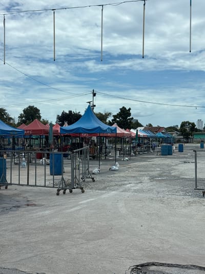 An outdoor market scene is depicted on a cloudy day in the city of Nonthaburi Municipality, Thailand. The market appears to be mostly empty, with numerous stalls set up under colorful tents. The tents are a mix of red, blue, and green. In front of the stalls are metal barricades on wheels. Several blue trash bins are visible amongst the stalls, along with white bags on the ground. The ground is paved with concrete, showing some signs of wear and tear, including cracks and puddles. In the background, trees and buildings can be seen. Overhead, wires are strung across the sky, with several vertical poles hanging down from them.