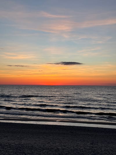 The image shows a sunset over the Baltic Sea in Garupe, Latvia. The sky is a mix of light blue, orange, and yellow hues, with wispy clouds and a darker cloud formation near the horizon. The sea is characterized by small waves reflecting light, separated by the sand and the border of the city on the right side of the image.