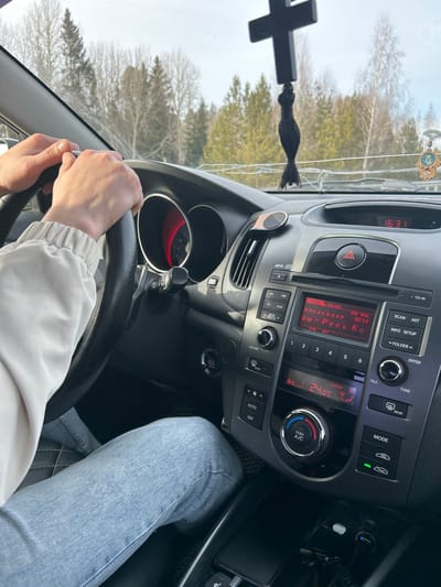 The image depicts the interior of a vehicle from the passenger perspective, looking towards the driver and through the windshield, situated in the city of Ir'yak, Russia.
**People:**
One individual is visible in the driver's seat. Their hands, bare, are positioned on the 10 and 2 o'clock positions of a black steering wheel, suggesting the vehicle is in operation or prepared to be. The driver wears a light-colored, possibly off-white, long-sleeved top with elasticized cuffs and light blue denim jeans. No facial features are visible.
**Vehicle Interior (Objects & Actions):**
The vehicle's dashboard is black. The driver-side instrument cluster features a circular design with a red backlight and a needle indicating a low or zero speed. The center console houses an automotive stereo system displaying Cyrillic text, specifically "Новая АЭС" (New NPP) and "Ио-Русь Бо," along with the time "16:31." Below the stereo, the climate control unit displays an internal temperature of "24.0°С" and "Outdoor" temperature. Controls for air conditioning, fan speed, and mode selection are present. A circular engine start/stop button is visible to the lower left of the center console. A black crucifix with a tassel hangs from the rearview mirror, and a blue and white Hamsa hand charm hangs on the passenger side of the dashboard or windshield. The windshield exhibits multiple horizontal cracks. In the center console storage area, a black smartphone and car keys are visible, along with a USB charger. The driver's footwell is covered by a black floor mat with a textured, possibly quilted, pattern.
**Environment (Exterior):**
Through the windshield, a line of trees is visible in the background, consisting of both evergreen conifers and bare deciduous trees, suggesting a late winter or early spring season. The sky is light, indicating daylight conditions.