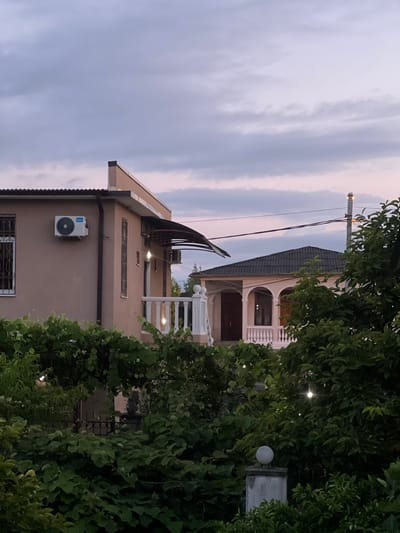The image depicts two houses in Ochamchyra, Abkhazia, under a cloudy dusk sky. The house in the foreground, painted a light tan, has a dark-tiled roof and an air conditioning unit mounted on its exterior wall. It features barred windows and a white-railed balcony with string lights. Lush green foliage, including grapevines, partially obscures the view of this house.
The second house, visible behind the first, has a dark-tiled roof and an arched entryway supported by columns. This house also has a white-railed balcony and is partially surrounded by trees. Overhead, power lines stretch across the sky, and a street lamp is visible on a pole. The overall scene is one of residential buildings in a vegetated area during twilight.