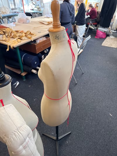 The image displays an interior view of a fashion or textile workshop, identified as being located in Arcueil, France. The primary subject in the immediate foreground is a beige fabric dress form mounted on a black metal tripod base, oriented slightly towards the left. This dress form features a wooden finial at the neck, which is marked with "95" and "L" (or "4"). Red string or tape is meticulously draped around the dress form, outlining contours from the neck, over the shoulders, down the back, and around the waist. To the lower left, a partial view of a second beige fabric dress form is visible, distinguished by red stitching delineating bust lines and what appears to be an attached white fabric element, possibly a sleeve or padded panel.
The floor throughout the visible area is covered with dark grey carpeting. In the midground to the left, a light-colored wooden work table with green metal legs is situated, laden with numerous cut fabric pieces, predominantly tan or gold in hue, alongside various workshop materials. Beneath this table, rolls of dark-colored fabric are stacked on shelves. The background reveals several individuals in various states of activity within the workshop: one person, seen from the back, wears a dark grey hoodie and dark pants; another individual with blonde hair, wearing a black top and light-colored pants, stands partially visible to the right of the first person; and a third person, dressed in a dark red top, is seated at a table further back on the right. A silver folding chair and a prominent bright pink plastic bag are also present in the background on the right side of the frame. The workshop space is brightly illuminated, suggesting ample ambient light.