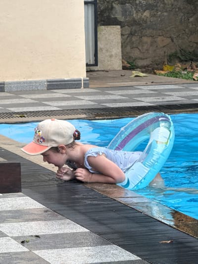 A young child wearing a light-colored hat and a blue-and-white outfit is partially submerged in a swimming pool. The child has a blue inner tube around their torso. The pool water is blue and partially obscures the child's body. The pool's edge is a tiled pattern of gray and white. There is an off-white wall and some greenery in the background. The image is slightly tilted to the side.