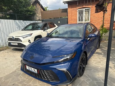 In Krtsanisi, Georgia, two Toyota cars are parked side-by-side on a paved surface. The car in the foreground is a dark blue sedan, possibly a Toyota Camry, with a black grille and black alloy wheels. Its license plate reads "1016 NL". The car behind it is a white SUV, likely a Toyota RAV4 Hybrid, with a black roof and black trim. Its license plate reads "SP-777-AM". The cars are parked in front of a brick building with barred windows. A tall, dark pole stands to the right of the blue sedan, and a black fabric canopy is draped over it. Trees and a wooden structure are visible in the background, along with a corrugated metal fence. The sky is partly cloudy.