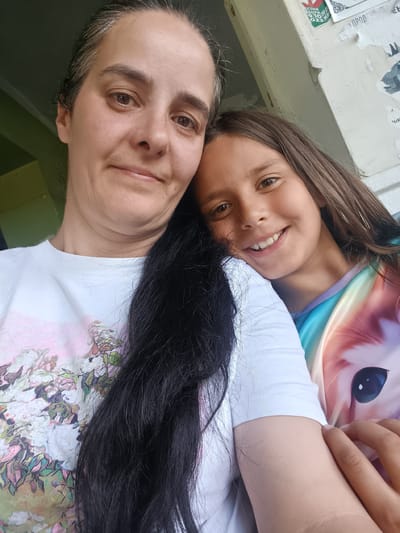 This media file is a close-up, tilted selfie featuring two people, likely a mother and daughter, in an indoor setting. The younger person is in the foreground, smiling warmly at the camera. She has brown eyes, brown hair, and is wearing a white t-shirt with a colorful graphic of a cat's face. The older person is behind and slightly above the younger person, also looking towards the camera with a gentle smile. She has brown eyes, dark hair with some grey streaks pulled back, and is wearing a white t-shirt with a floral pattern. Long, dark hair from the older person drapes over the younger person's shoulder and chest. The background shows hints of a wall with peeling posters, suggesting a somewhat aged or utilitarian environment. Some visible text on the posters appears to be in Cyrillic script, with one poster having green, white, and red colors, possibly representing the Bulgarian flag. The lighting suggests it could be daytime. The overall mood is affectionate and casual.